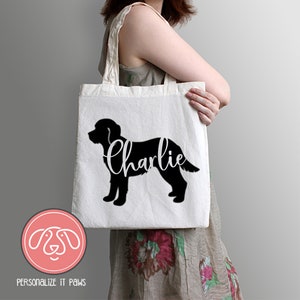 May include: A white canvas tote bag featuring a black silhouette of a dog with the name "Charlie" in cursive script. The bag also has a circular logo with a dog's face and the text "PERSONALIZE IT PAWS".