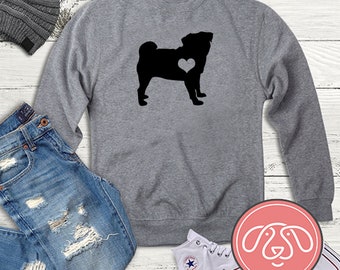 Könnte beinhalten: Grauer Pullover mit einer schwarzen Silhouette eines Mops mit einem Herzen auf der Brust. Der Pullover liegt auf einer Holzoberfläche mit einer Jeans, einer Mütze und einem Paar weißen Turnschuhen.
