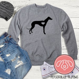 K&ouml;nnte beinhalten: Ein graues Sweatshirt mit einer schwarzen Silhouette eines Windhundes mit einem Herzen in der Brust. Das Sweatshirt liegt auf einer wei&szlig;en Holzoberfl&auml;che mit einer blauen Jeans, einer grauen M&uuml;tze und wei&szlig;en Turnschuhen.