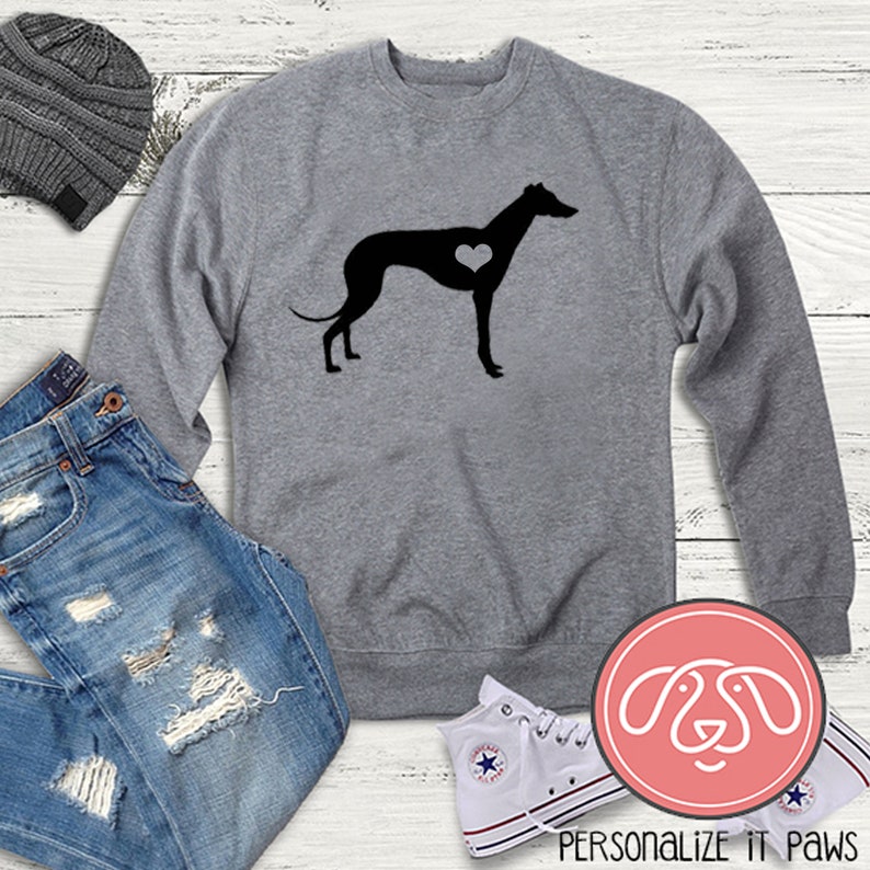 K&ouml;nnte beinhalten: Ein graues Sweatshirt mit einer schwarzen Silhouette eines Windhundes mit einem Herzen in der Brust. Das Sweatshirt liegt auf einer wei&szlig;en Holzoberfl&auml;che mit einer blauen Jeans, einer grauen M&uuml;tze und wei&szlig;en Turnschuhen.