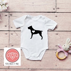 May include: White baby onesie with a black silhouette of a Cane Corso dog with a white heart on its chest. The onesie has short sleeves and a snap closure. The background is a light wood surface.