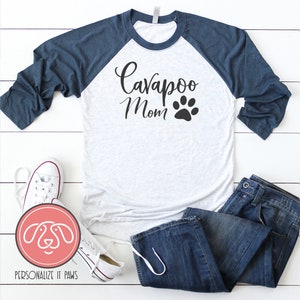 May include: A white and navy blue baseball tee shirt with the text "Cavapoo Mom" and a black paw print. The shirt is laying on a white wooden surface with a pair of white sneakers, blue jeans, and a pink and white logo.