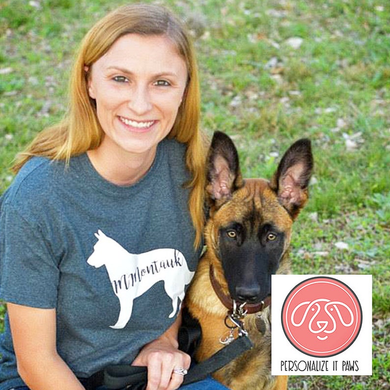 May include: A woman wearing a teal blue t-shirt with a white silhouette of a dog and the text "Montauk" on it. She is sitting on the grass with a brown and black German Shepherd dog. The dog is wearing a brown leather collar and a leash. A pink circle with a white outline and a stylized dog silhouette inside is in the lower right corner of the image. The text "PERSONALIZE IT PAWS" is below the circle.