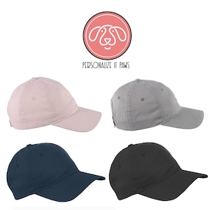 May include: Four blank baseball caps in different colors: pink, gray, navy blue, and black. The caps are all the same style, with a curved brim and a small adjustable strap in the back. The caps are arranged in a grid pattern on a white background.  Personalize It Paws.