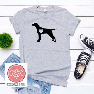 Puede incluir: Una camiseta gris con una silueta negra de un perro con un corazón en el pecho. El texto "PERSONALIZE IT PAWS" está en un círculo rosa debajo de la imagen.