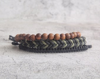 May include: A stack of three bracelets. The top bracelet features smooth, round, wooden beads in a warm brown hue. Below it is a braided bracelet with black and olive green cord. The bottom bracelet is a solid black, woven design.