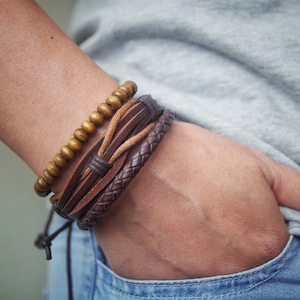 Könnte beinhalten: Ein Set aus drei Armbändern an einem Handgelenk. Die Armbänder sind aus braunem Leder und Holzperlen.