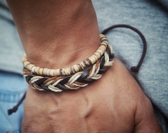 May include: A braided leather bracelet with brown, white, and beige strands, layered over a bracelet made of small, light brown beads.