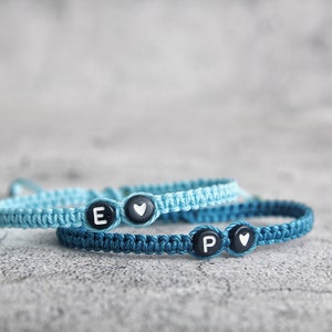 May include: Two friendship bracelets with blue and light blue cords. One bracelet has a black bead with the letter "E" and a heart, the other has a black bead with the letter "P" and a heart.