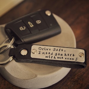 Könnte beinhalten: Ein schwarzer Leder-Schlüsselanhänger mit einem silbernen Metalletikett, auf dem "Drive Safe. I need you here with me xoxo J" steht.