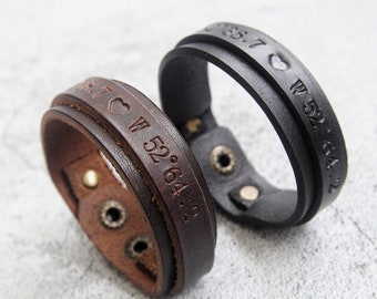 May include: Two leather bracelets with engraved coordinates and a heart symbol. The bracelet on the left is brown and the bracelet on the right is black.