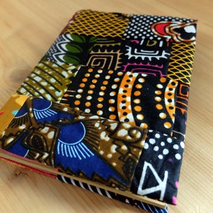 Peut inclure: Un journal fait à la main avec une couverture patchwork. La couverture présente une variété de tissus africains imprimés à la cire colorés dans des tons de bleu, jaune, vert et noir. Le journal est rectangulaire et semble fermé.