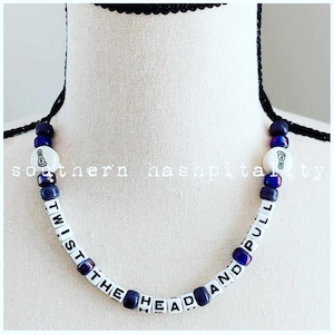 May include: A white beaded necklace with black lettering spelling "Twist the head and pull". The beads are interspersed with small blue and purple beads. The necklace is on a white mannequin.