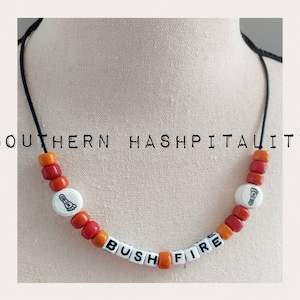 May include: A necklace made of orange and red beads with white beads that spell out "BUSH FIRE" and have a footprint design. The necklace is on a black cord and the text "SOUTHERN HOSPITALITY" is written above the necklace.