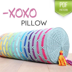May include: A colourful crochet pillow with a rainbow pattern and a grey background. The pillow has a tassel on one end and says "XOXO Pillow" on the front.