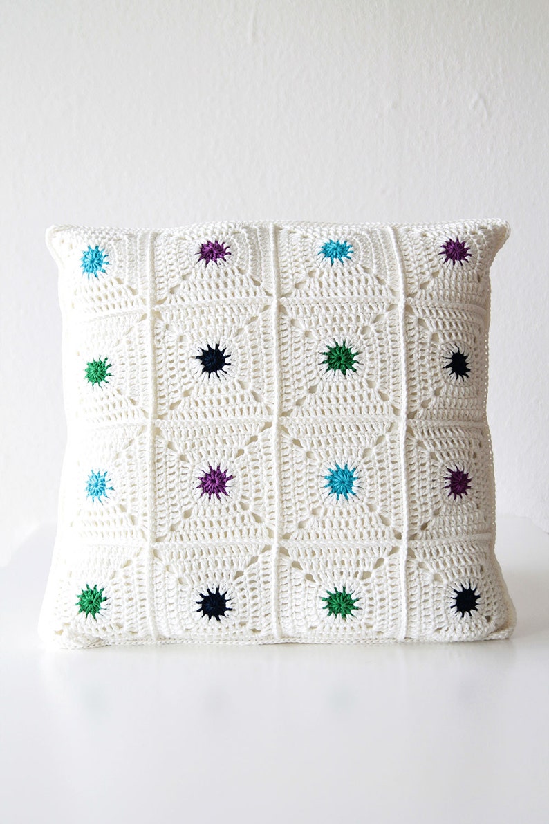 May include: White crocheted pillow with a geometric pattern and colorful accents. The pillow is decorated with small, colorful circles in shades of blue, green, purple, and black.
