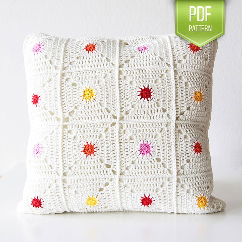 May include: White crochet pillow cover with a geometric pattern and colorful star accents. The pillow cover is made with a white yarn and features a repeating square pattern with a star in the center of each square. The stars are in various colors, including red, pink, yellow, and orange.
