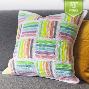 Può includere: Un cuscino bianco all'uncinetto con un motivo a righe colorate. Il cuscino è realizzato con una varietà di colori, tra cui verde, blu, rosa e giallo. Il cuscino è seduto su un divano grigio.