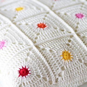 May include: White crocheted throw pillow with colorful embroidered flowers. The pillow is made of a soft, textured fabric and features a variety of colors, including red, pink, yellow, and orange.
