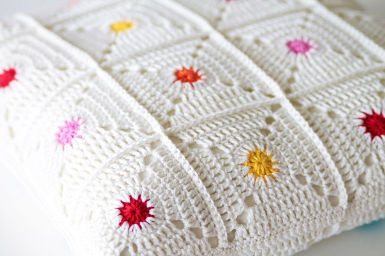 May include: White crocheted throw pillow with colorful embroidered flowers. The pillow is made of a soft, textured fabric and features a variety of colors, including red, pink, yellow, and orange.
