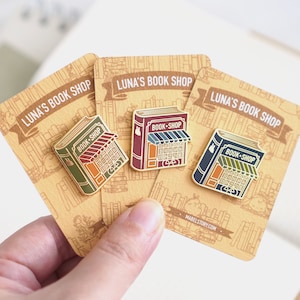 May include: Three enamel pins shaped like books with the text "Book-Shop" on the front. The pins are in different colors: green, red, and blue. The pins are on cards with the text "Luna's Book Shop" on them.