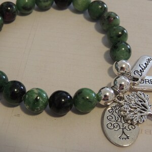 May include: A green gemstone bracelet with silver beads and charms. The charms include a tree of life design, and two tags that say "Believe" and "Dream".