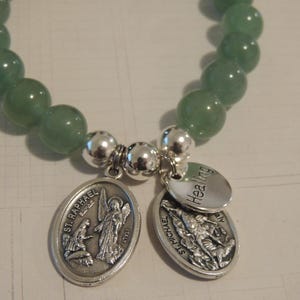 May include: A green gemstone bracelet with silver charms. The charms feature the words "Healing" and "St. Raphael" with images of angels.