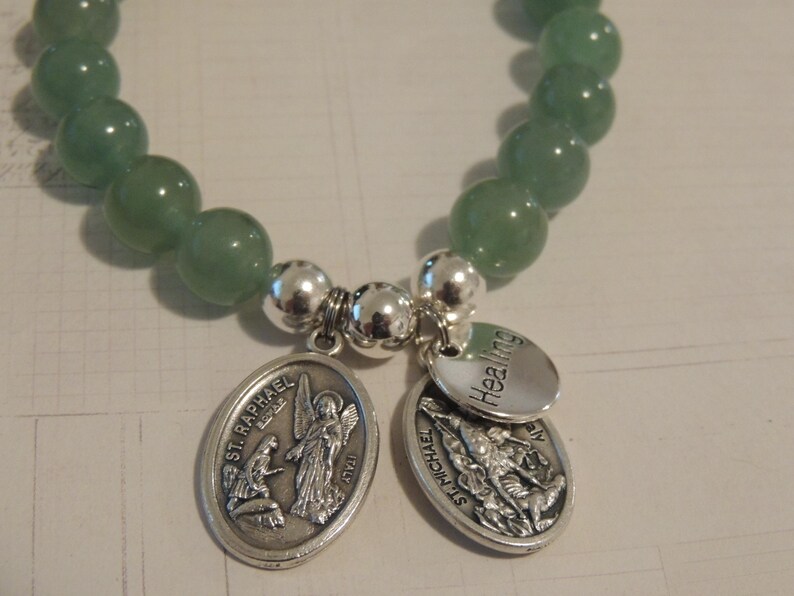 May include: A green gemstone bracelet with silver charms. The charms feature the words "Healing" and "St. Raphael" with images of angels.