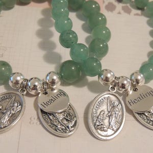 May include: Two green stone beaded bracelets with silver charms. One charm features a depiction of Saint Raphael and the text "Healing". The other charm features a depiction of Saint Michael and the text "Healing".