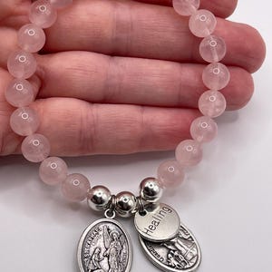 May include: A rose quartz beaded bracelet with two silver charms. One charm is oval-shaped with an image of St. Raphael and the text "ST. RAPHAEL". The other charm is round and says "Healing".