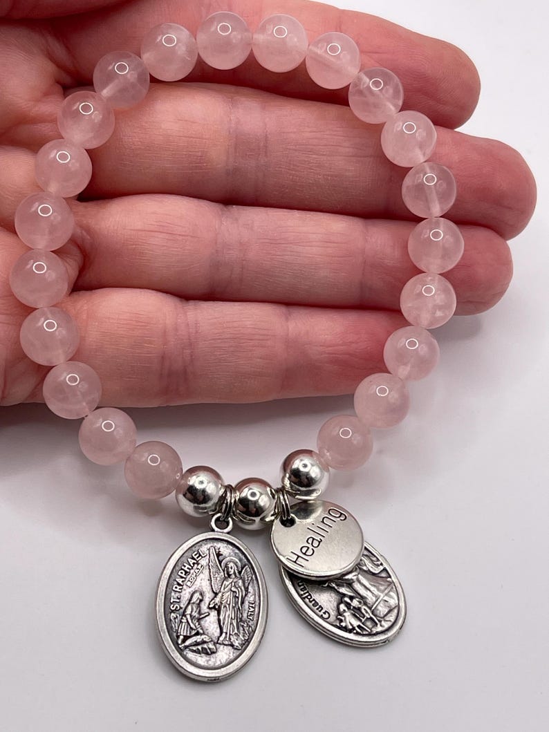 May include: A rose quartz beaded bracelet with two silver charms. One charm is oval-shaped with an image of St. Raphael and the text "ST. RAPHAEL". The other charm is round and says "Healing".