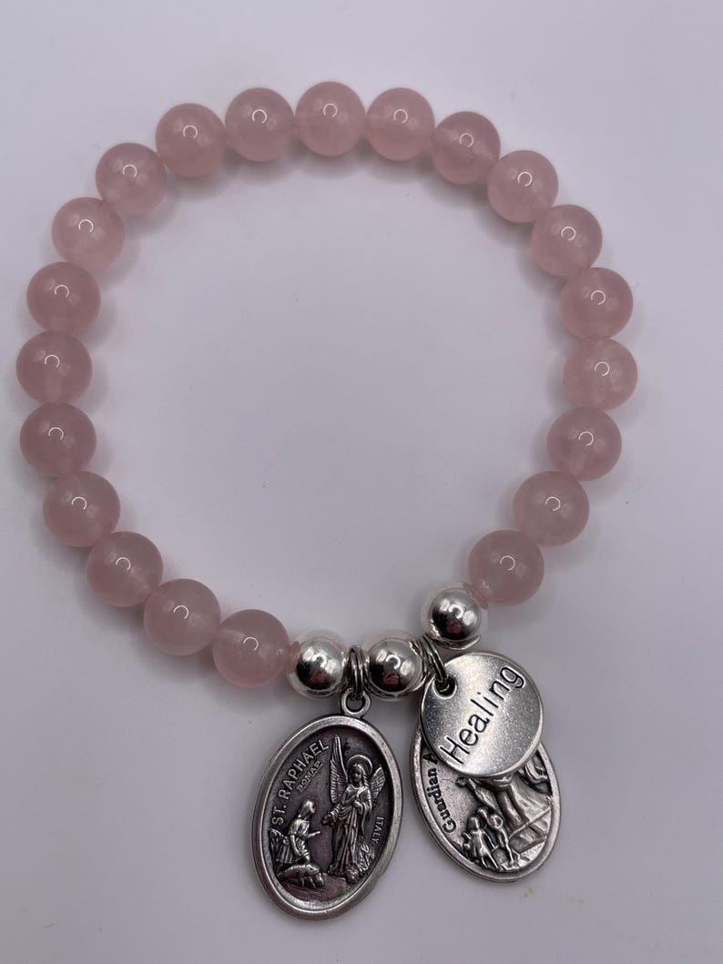 May include: A rose quartz beaded bracelet with two silver charms. One charm features an image of St. Raphael, and the other says "Healing". The bracelet is made of round, light pink beads.