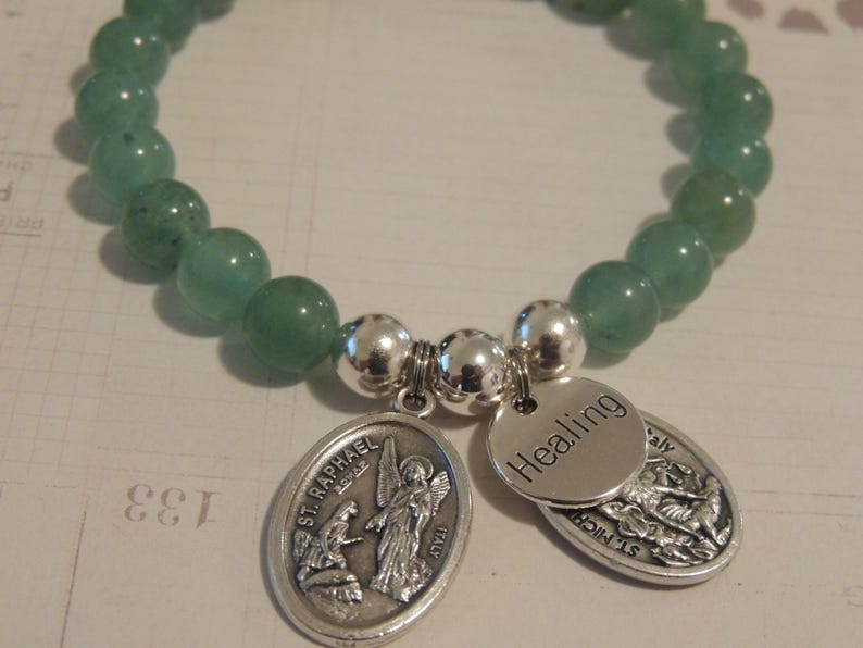 May include: A green gemstone bracelet with silver beads and charms. The charms feature the words "Healing" and "St. Raphael" with religious imagery.