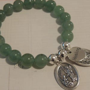 May include: A green gemstone bracelet with silver beads and two charms. One charm is a silver oval with the words "St. Raphael" and an image of an angel. The other charm is a silver oval with the word "Healing".