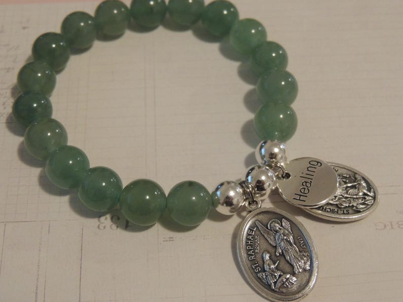 May include: A green gemstone bracelet with silver beads and two charms. One charm is a silver oval with the words "St. Raphael" and an image of an angel. The other charm is a silver oval with the word "Healing".