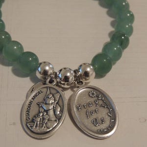May include: A green gemstone bracelet with two silver charms. One charm features a guardian angel with the words "Guardian Angel" and the other charm says "Pray for Us".