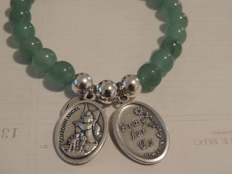 May include: A green gemstone bracelet with two silver charms. One charm features a guardian angel with the words "Guardian Angel" and the other charm says "Pray for Us".