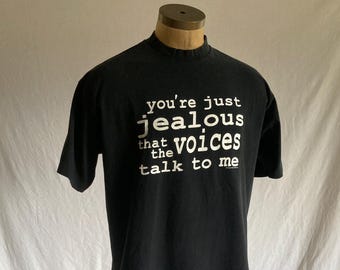 May include: Black t-shirt with the white text "you're just jealous that the voices talk to me." The shirt has short sleeves and a crew neck. The shirt is displayed on a mannequin with light green pants.