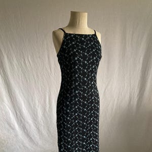 Puede incluir: Un vestido negro con escote cuadrado y tirantes finos. El vestido está adornado con un delicado estampado floral en un color más claro. El vestido se exhibe en un maniquí.