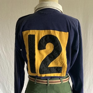 May include: Navy blue long-sleeved shirt with a white collar and a yellow patch on the back displaying the number 12 in black. The shirt is worn with olive green trousers and a striped belt.