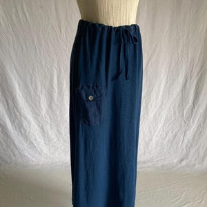May include: A long, navy blue skirt with an elastic waistband and drawstring. The skirt features a single pocket on the front with a button closure. The fabric appears to be a soft, flowing material.