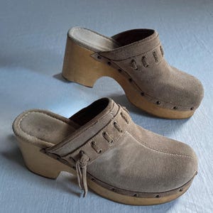 May include: A pair of light brown suede clogs with wooden soles and heels. The clogs have a slip-on design with decorative stitching and small metal studs around the sole. The heels are approximately 3 inches high.