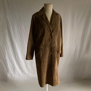 May include: A brown suede coat displayed on a mannequin. The coat has a classic design with a notched collar, three buttons, and two front pockets. The coat is knee-length and has long sleeves.