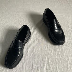 May include: A pair of black leather penny loafers. These classic shoes feature a rounded toe, a strap across the vamp, and a low heel. The leather has a glossy finish. The shoes are displayed on a white surface.