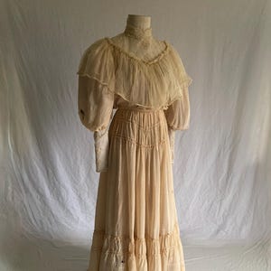 May include: A vintage cream-colored dress with a high neckline and long sleeves. The dress features a ruffled bodice, a cinched waist, and a tiered skirt. The dress is displayed on a mannequin and paired with black boots.