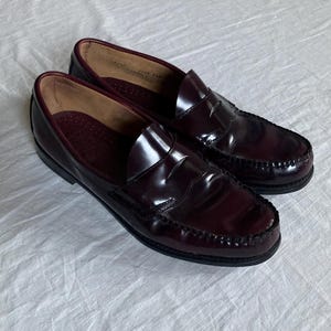 May include: A pair of burgundy leather penny loafers. The shoes have a glossy finish, a stitched vamp, and a dark sole. The interior is a lighter shade of burgundy. The shoes are displayed on a white surface.