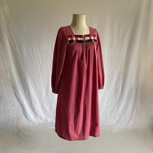 May include: A dusty rose-colored dress with long sleeves and a square neckline. The neckline is accented with a decorative band of black, white, and red plaid. The dress is displayed on a mannequin against a white backdrop.