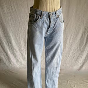 May include: Light blue denim jeans featuring a classic five-pocket design, a button fly closure, and a straight-leg cut. The denim has a faded wash with subtle whiskering details at the hips and a standard waistband with belt loops.