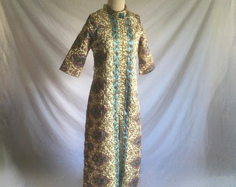 May include: A vintage cream-colored dress with a high collar and three-quarter sleeves. The dress features a repeating paisley pattern in shades of burgundy, teal, and gold. Teal trim runs down the front and around the collar. The dress is displayed on a mannequin.