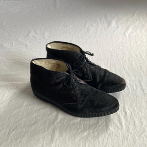 May include: A pair of black ankle boots with a furry texture and a lace-up closure. The boots have a white lining and a black rubber sole.